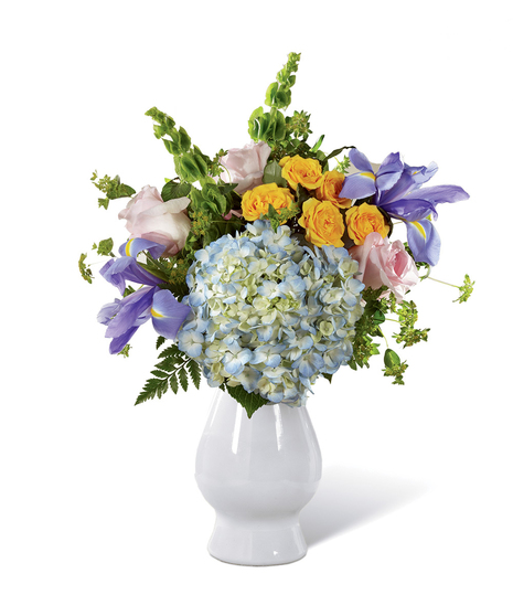 Pink roses, yellow spray roses, stalks of green Bells of Ireland, blue iris, clouds of blue hydrangea, bupleurum, and lush greens in a pale blue ceramic vase.