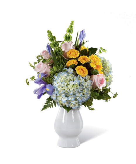 Pink roses, yellow spray roses, stalks of green Bells of Ireland, blue iris, clouds of blue hydrangea, bupleurum, and lush greens in a pale blue ceramic vase.