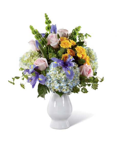 Pink roses, yellow spray roses, stalks of green Bells of Ireland, blue iris, clouds of blue hydrangea, bupleurum, and lush greens in a pale blue ceramic vase.